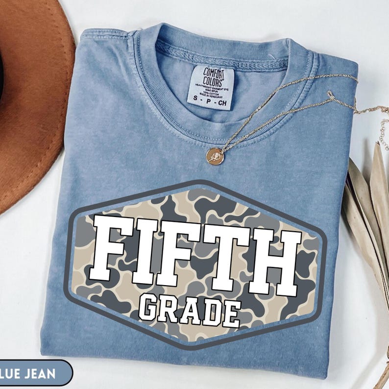 Pu&ograve; includere: Una t-shirt blu a maniche corte con scollo rotondo ed etichetta Comfort Colors. Al centro &egrave; presente una grafica esagonale con motivo mimetico beige e grigio, contenente il testo bianco FIFTH GRADE con un contorno nero.