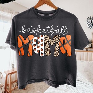 May include: Dark gray t-shirt with the words "basketball MAMA" in white script and a basketball-themed design. The word "MAMA" features basketballs, leopard print, and basketball textures. A casual, comfortable tee.