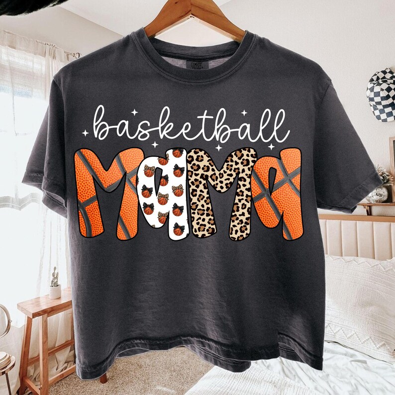 May include: Dark gray t-shirt with the words "basketball MAMA" in white script and a basketball-themed design. The word "MAMA" features basketballs, leopard print, and basketball textures. A casual, comfortable tee.