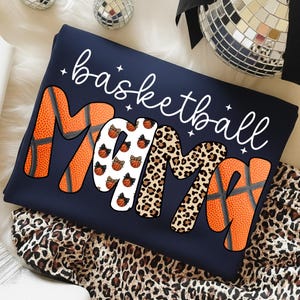 May include: Navy blue sweatshirt with the words "basketball Mama" in white script. The letters "M" and "A" are orange basketballs, the second "M" is leopard print, and the middle "M" has basketballs with bows.