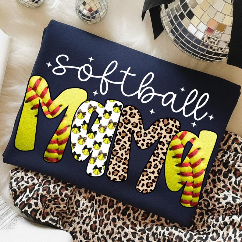 May include: Navy blue sweatshirt with the words "softball mama" in a playful design. The word "softball" is in white script, and "mama" is in a mix of softball and leopard print patterns. The sweatshirt is folded on a leopard print fabric.