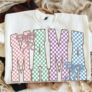 Pode incluir: Camisola branca com a palavra "MAMA" em letras grandes e coloridas em xadrez. As letras são rosa, verde, roxo e azul, com contorno de estampa de leopardo. Laços rosa e azuis também são apresentados.