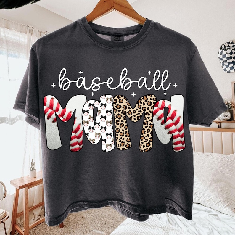 May include: Dark gray t-shirt with the words "baseball Mama" in white script and decorative lettering. The word "baseball" is in script, and the word "Mama" features baseball stitching, bows, and leopard print.