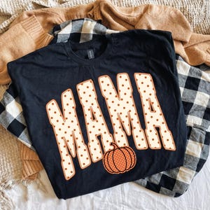 May include: A black short-sleeved t-shirt featuring the word MAMA in large, cream-colored block letters with a brown polka dot pattern. An orange pumpkin with a matching brown polka dot design is positioned below the letter A.
