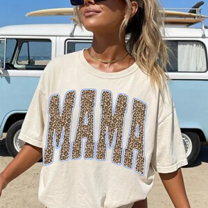May include: A cream-colored short-sleeve t-shirt featuring the word MAMA printed in a large, bold, collegiate-style font. The letters have a brown and black leopard print pattern and are outlined with a thin, vibrant blue border.