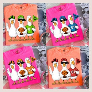 May include: Four brightly coloured t-shirts, each featuring a cartoon image of three ducks wearing hats and holding a basketball. The ducks are wearing jerseys with the number "01" and holding a "GO TEAM" flag. The shirts are in shades of pink and orange.