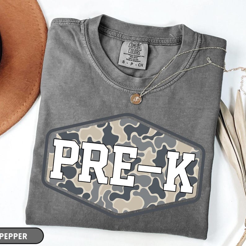 Pu&ograve; includere: Una t-shirt a maniche corte grigio antracite con una grafica esagonale caratterizzata da un motivo mimetico nei toni del grigio, beige e nero. Il testo PRE-K &egrave; stampato in grandi lettere bianche in stampatello al centro dell'esagono.