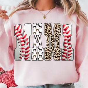 May include: A light pink sweatshirt with the word "MAMA" in large, bold letters. The letters have unique patterns: baseball stitching, bows, and leopard print. The sweatshirt is a casual, comfortable style.