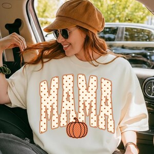 May include: A cream-colored crewneck sweatshirt featuring the word MAMA in large, block-style letters with a brown polka dot pattern. An orange pumpkin graphic with a matching brown polka dot pattern is positioned below the text.
