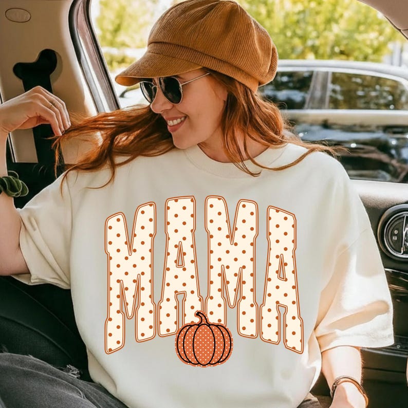 May include: A cream-colored crewneck sweatshirt featuring the word MAMA in large, block-style letters with a brown polka dot pattern. An orange pumpkin graphic with a matching brown polka dot pattern is positioned below the text.