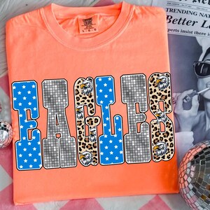 May include: A coral-colored t-shirt with the word "EAGLES" in large, bold letters. The letters have a mix of designs: blue with white stars, a disco ball pattern, and leopard print with eagle heads. The shirt has a crew neck.