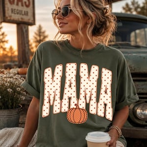 May include: An olive green short-sleeve t-shirt featuring the word MAMA in large, cream-colored block letters with orange polka dots. An orange pumpkin with cream-colored polka dots is positioned at the base of the letter M in the center.