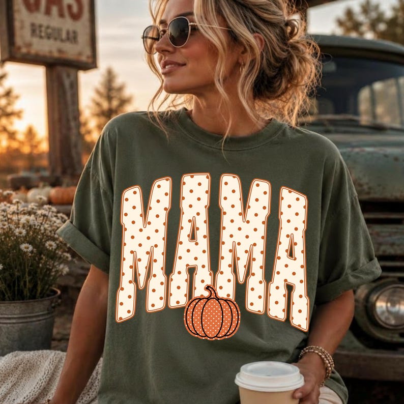 May include: An olive green short-sleeve t-shirt featuring the word MAMA in large, cream-colored block letters with orange polka dots. An orange pumpkin with cream-colored polka dots is positioned at the base of the letter M in the center.
