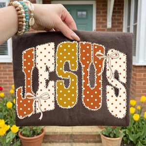 Op de afbeelding: Een donkerbruin t-shirt met het woord JESUS in grote, blokvormige letters. De letters hebben een wit en oranje stippenpatroon met een luipaardprint rand. Twee lichtroze lintstrikjes zijn bevestigd aan de letters J en S.