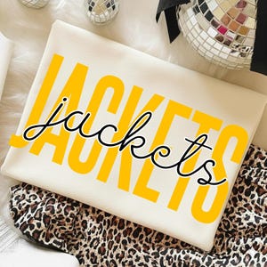 Puede incluir: Sudadera crema con la palabra "JACKETS" en letras amarillas en negrita, superpuesta con la palabra "jackets" en cursiva negra. La sudadera está doblada sobre una tela con estampado de leopardo.
