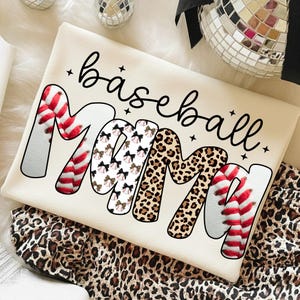 May include: Cream-colored baseball Mama t-shirt. The word "Mama" is designed with baseball stitching, bows, and leopard print. The word "baseball" is written in a cursive font. The shirt is folded on a leopard print fabric.