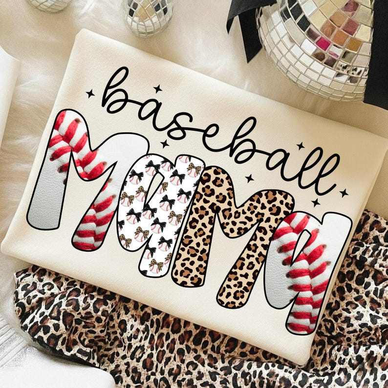 May include: Cream-colored baseball Mama t-shirt. The word "Mama" is designed with baseball stitching, bows, and leopard print. The word "baseball" is written in a cursive font. The shirt is folded on a leopard print fabric.