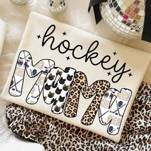 May include: Cream-colored sweatshirt with the words "hockey mom" in black script. The letters of "MOM" are decorated with a leopard print, black bows, and hockey-themed graphics. The sweatshirt is on a leopard print fabric.