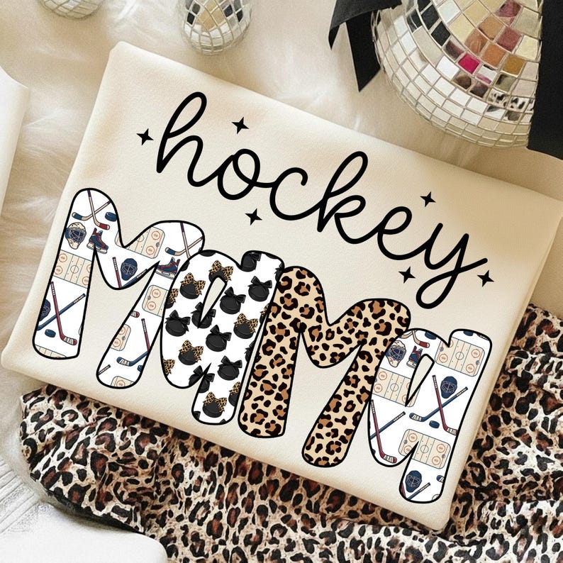 May include: Cream-colored sweatshirt with the words "hockey mom" in black script. The letters of "MOM" are decorated with a leopard print, black bows, and hockey-themed graphics. The sweatshirt is on a leopard print fabric.