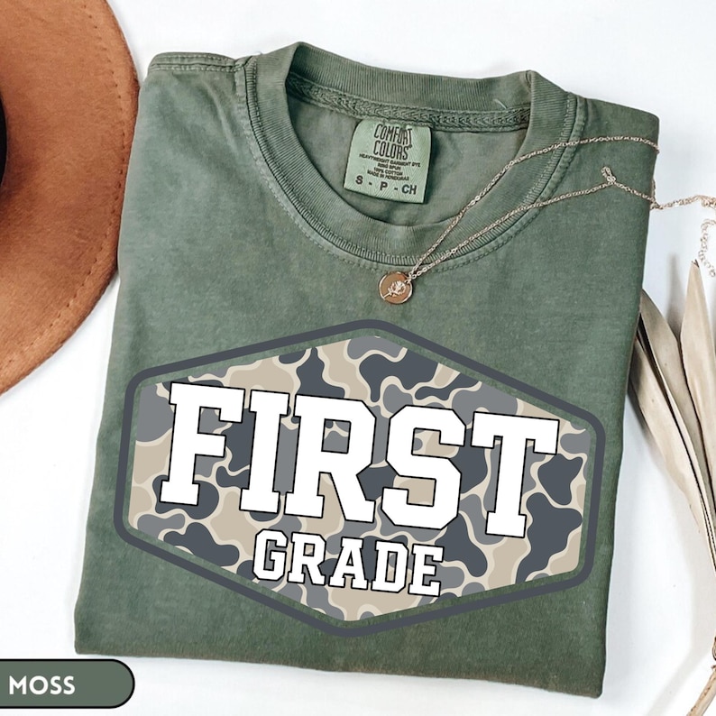 Pu&ograve; includere: Una t-shirt a maniche corte color verde muschio con scollo rotondo. Al centro presenta una grafica esagonale con un motivo mimetico beige, grigio e grigio scuro. Il testo FIRST GRADE &egrave; stampato in grassetto bianco sulla grafica.