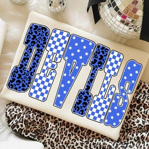 Puede incluir: Sudadera de color crema con la palabra "DEVILS" en una fuente audaz y estilizada. Las letras presentan estampado de leopardo azul, patrones de cuadros y estrellas blancas. La sudadera se muestra sobre una tela con estampado de leopardo.