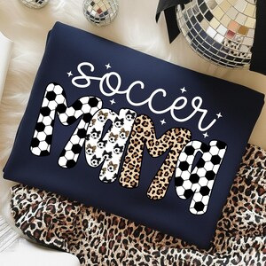 May include: Navy blue sweatshirt with the words "Soccer Mama" in a playful design. The word "Mama" features a mix of soccer ball, leopard print, and soccer ball print patterns. The word "Soccer" is in white script.