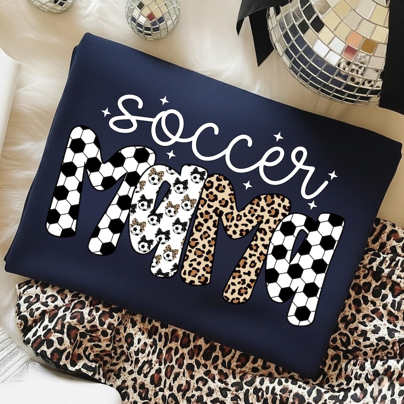 May include: Navy blue sweatshirt with the words "Soccer Mama" in a playful design. The word "Mama" features a mix of soccer ball, leopard print, and soccer ball print patterns. The word "Soccer" is in white script.