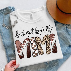 May include: White t-shirt with the words "football MIMI" in black script and decorative fonts. The letters "MIMI" are designed with footballs and leopard print. The shirt is displayed on a pair of blue jeans.