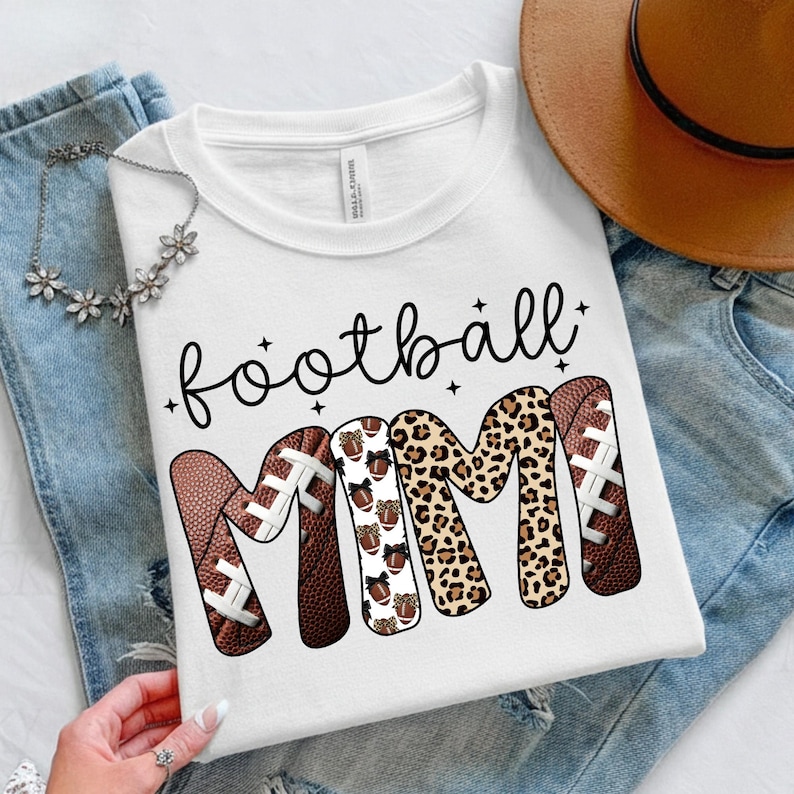 May include: White t-shirt with the words "football MIMI" in black script and decorative fonts. The letters "MIMI" are designed with footballs and leopard print. The shirt is displayed on a pair of blue jeans.