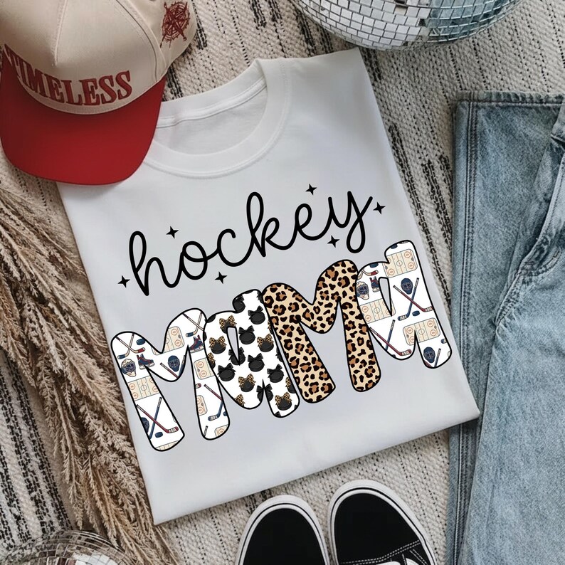 May include: White t-shirt with the words "hockey mom" in black script and patterned letters. The letter "M" is decorated with leopard print and cat faces. Hockey sticks and pucks decorate the other letters. A red and tan baseball cap is in the background.