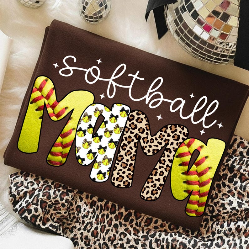 May include: A brown t-shirt with the words "softball mama" in white script. The word "mama" is designed with softball and leopard print patterns. The shirt is folded and placed on a leopard print fabric.