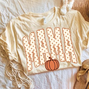 May include: A cream-colored short-sleeve t-shirt featuring the word MAMA in large, block-style letters. The letters are white with brown polka dots and a brown outline. A small, orange pumpkin with white polka dots is positioned below the text.