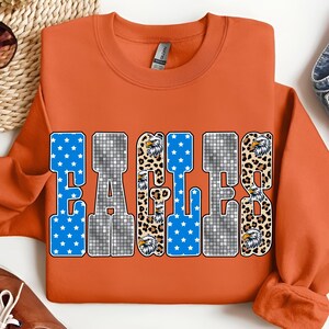 May include: An orange sweatshirt with the word "EAGLES" in a bold, block letter design. The letters feature a mix of blue stars, silver mosaic, and leopard print with eagle head graphics. A casual, sporty apparel item.
