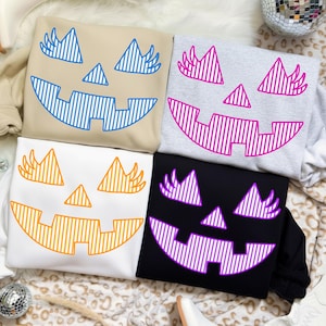 May include: Four sweatshirts with jack-o'-lantern face designs. The sweatshirts are beige, gray, white, and black. Each has a different color for the face design: blue, pink, orange, and purple. The designs feature triangle eyes, a triangle nose, and a smiling mouth.