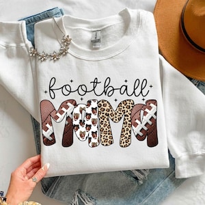 May include: White sweatshirt with the words "football mom" in black script. The letters "MOM" are designed with football patterns and leopard print. A floral necklace and a brown hat are also visible.
