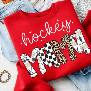 May include: A red sweatshirt with the words "hockey mama" in white script and block letters. The word "mama" features a mix of patterns, including hockey-themed graphics, leopard print, and black cat silhouettes.