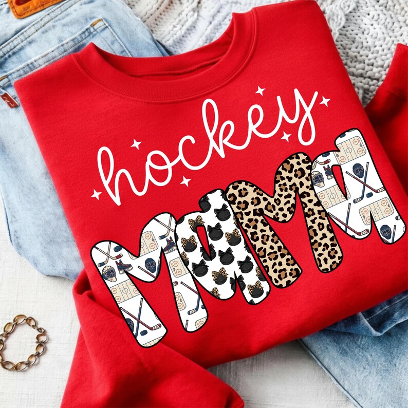 May include: A red sweatshirt with the words "hockey mama" in white script and block letters. The word "mama" features a mix of patterns, including hockey-themed graphics, leopard print, and black cat silhouettes.