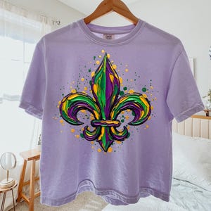May include: A lilac t-shirt with a colourful fleur-de-lis emblem in green, yellow, and purple. The design has a textured, painted look with paint splatters. The t-shirt is displayed on a wooden hanger.