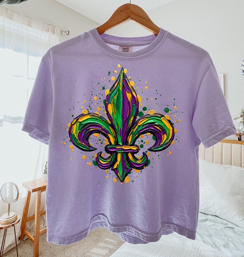 May include: A lilac t-shirt with a colourful fleur-de-lis emblem in green, yellow, and purple. The design has a textured, painted look with paint splatters. The t-shirt is displayed on a wooden hanger.