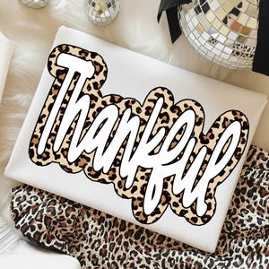 Peut inclure: Un coussin rectangulaire blanc avec le mot "Thankful" en gras et en cursive. Le texte est bord&eacute; d'un motif l&eacute;opard dans les tons marron et beige. Le coussin est pos&eacute; sur un tissu &agrave; imprim&eacute; l&eacute;opard.