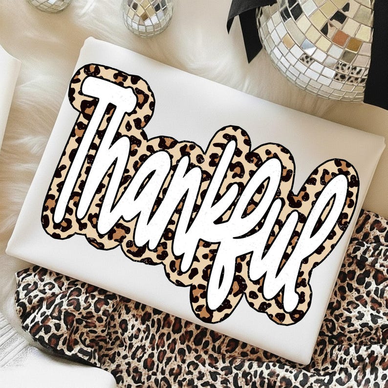 Peut inclure: Un coussin rectangulaire blanc avec le mot "Thankful" en gras et en cursive. Le texte est bord&eacute; d'un motif l&eacute;opard dans les tons marron et beige. Le coussin est pos&eacute; sur un tissu &agrave; imprim&eacute; l&eacute;opard.