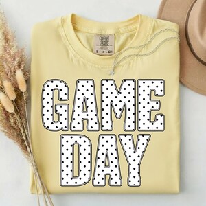 May include: A light yellow t-shirt with the words "GAME DAY" in large, black-outlined letters filled with white polka dots. A silver necklace with a star pendant and a tan hat are visible.