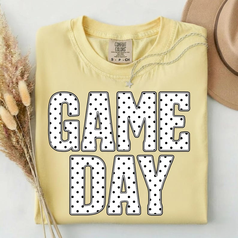 May include: A light yellow t-shirt with the words "GAME DAY" in large, black-outlined letters filled with white polka dots. A silver necklace with a star pendant and a tan hat are visible.