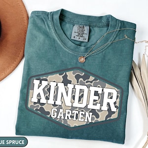 Pu&ograve; includere: Una t-shirt color blu abete con una grafica esagonale caratterizzata da un motivo mimetico beige e grigio. Il testo "KINDER" appare in grandi lettere bianche sopra la parola "GARTEN" scritta in lettere bianche pi&ugrave; piccole all'interno.