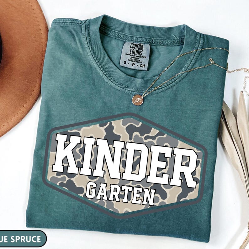 Pu&ograve; includere: Una t-shirt color blu abete con una grafica esagonale caratterizzata da un motivo mimetico beige e grigio. Il testo "KINDER" appare in grandi lettere bianche sopra la parola "GARTEN" scritta in lettere bianche pi&ugrave; piccole all'interno.