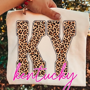 May include: A cream-coloured t-shirt featuring the large block letters KY filled with a brown and black leopard print pattern. Below the letters, the word Kentucky is printed in a bright pink cursive script font with a dark brown outline.