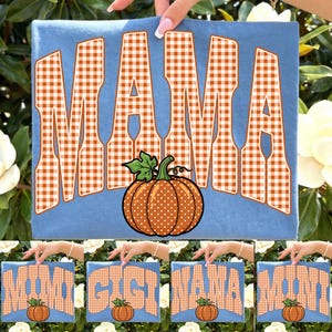 Puede incluir: Una camiseta azul claro con la palabra MAMA en letras grandes de cuadros vichy naranjas y blancos. Debajo del texto hay una calabaza naranja redonda con lunares blancos y un tallo verde. El diseño está impreso en la tela.
