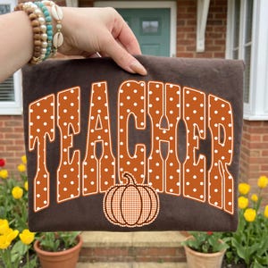 May include: A dark brown t-shirt featuring the word TEACHER in large, orange, white-polka-dotted letters. Below the text is a graphic of an orange pumpkin with a white checkered pattern. The shirt is held up against a blurred outdoor background.