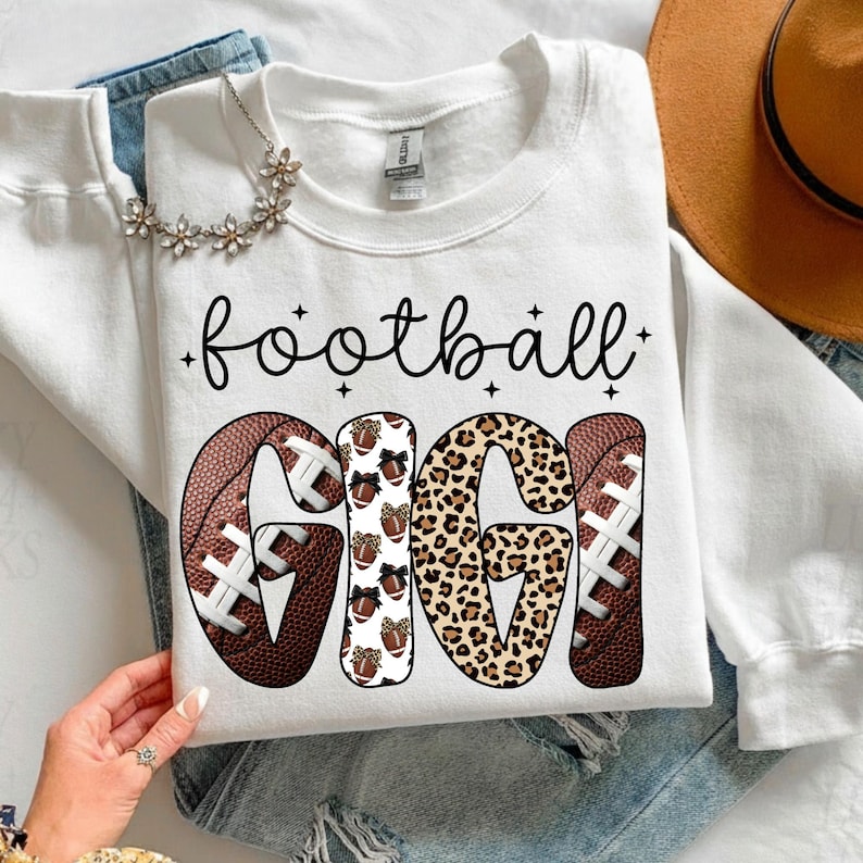 May include: White sweatshirt with the words "football GIGI" in black script and bold letters. The letters "GIGI" are decorated with footballs, leopard print, and footballs with bows. A floral necklace and a brown hat are visible.