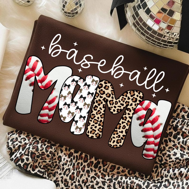 May include: A brown t-shirt with the words "baseball Mama" in white script and bold letters. The letter "M" is designed with baseball stitching and leopard print. The shirt is folded and placed on a leopard print fabric.